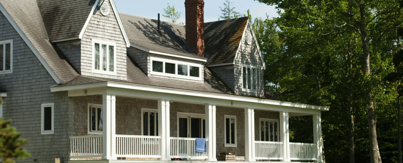 Gray shingle house with white porch, chimney, and green lawn surrounded by trees on a sunny day