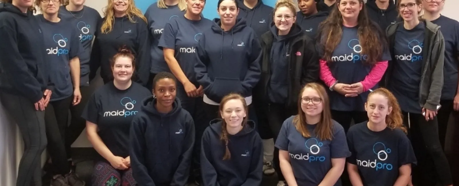 Group photo of MaidPro cleaning service team wearing branded shirts in office with blue wall art.