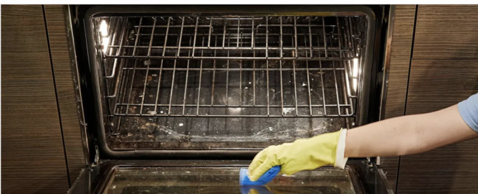 Person wearing yellow gloves cleaning a dirty oven door with a blue sponge in a modern kitchen.