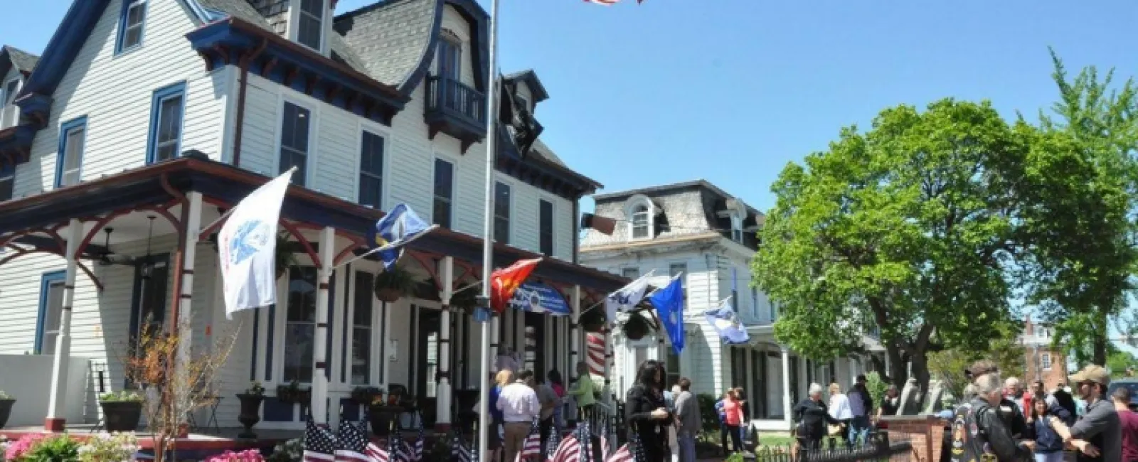 Crowd gathered around historic house decorated with American flags under clear blue sky on a sunny day