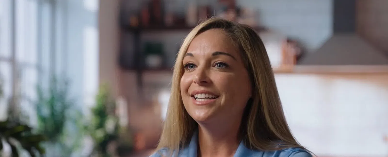 Smiling woman with blonde hair wearing a blue shirt in a bright, blurred kitchen setting.