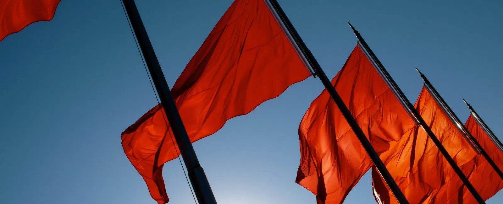 Bright red flags waving on poles against a clear blue sky with sunlight behind them