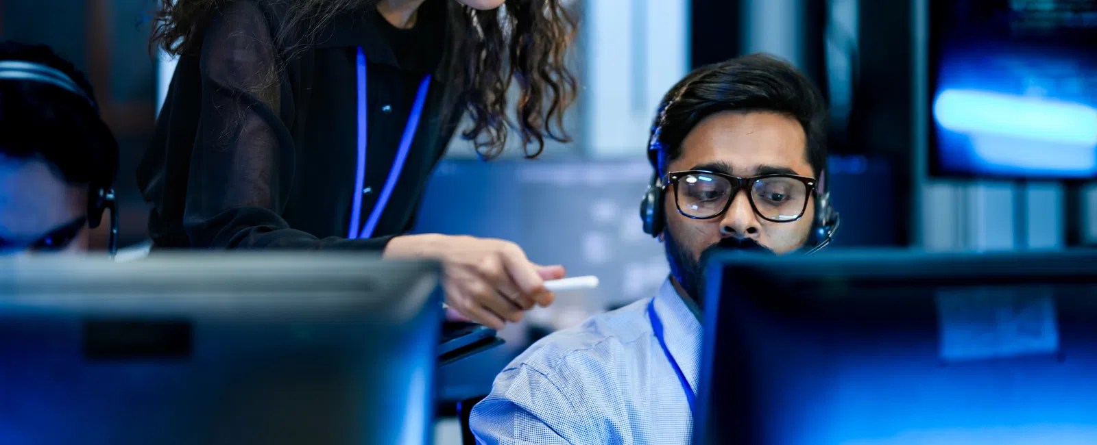 Female technician assists male colleague at computer in a modern control room during nighttime shift.
