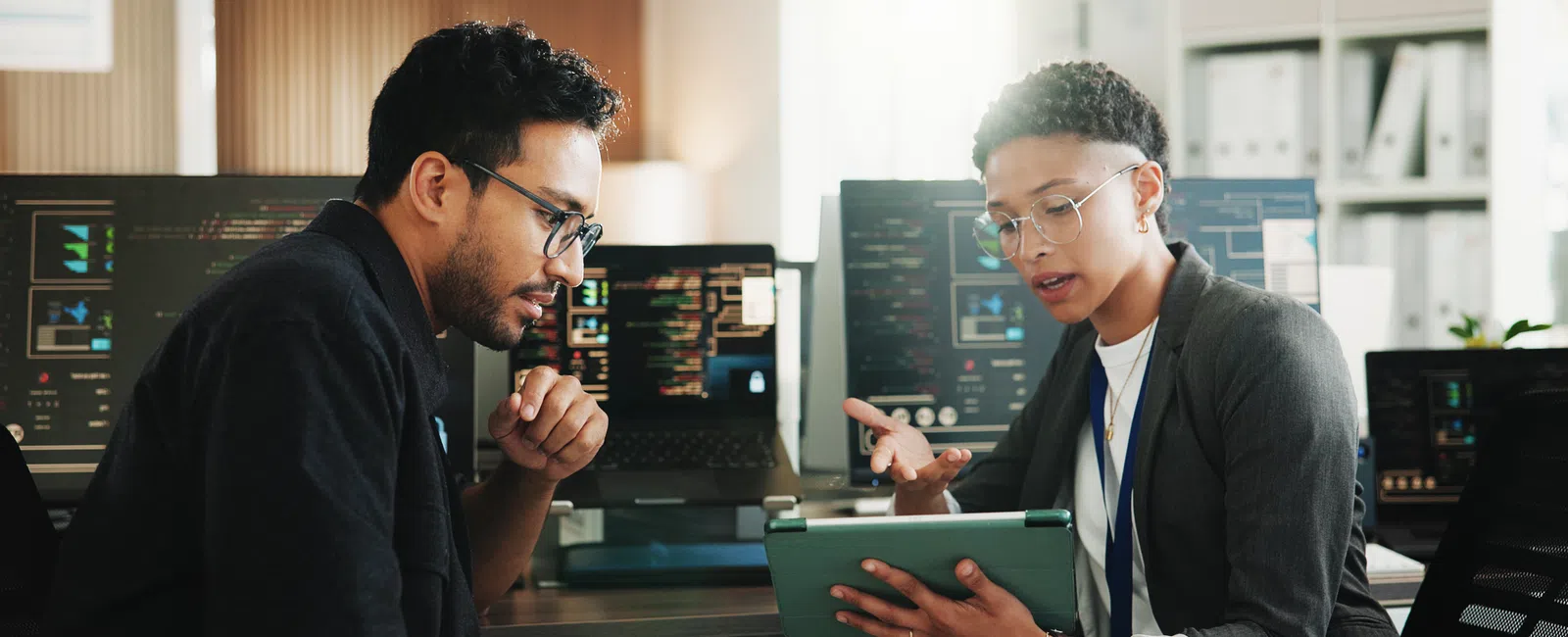 Two professionals discuss software development while reviewing code on a tablet in a modern office setting.