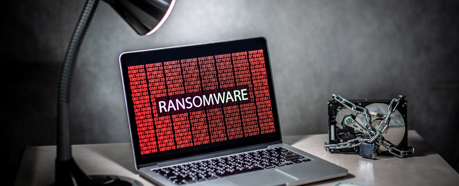 Laptop screen displaying ransomware warning with binary code, desk lamp, locked hard disk, and money on table.