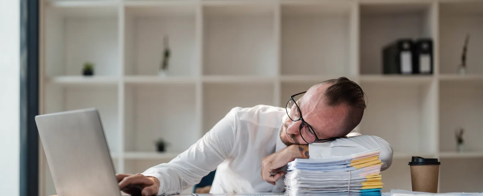 Tired man resting head on tall stack of documents while working on laptop in office.