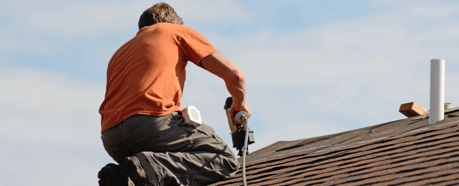 Roofer in orange shirt kneeling on shingled roof using nail gun under a blue sky during roof installation.