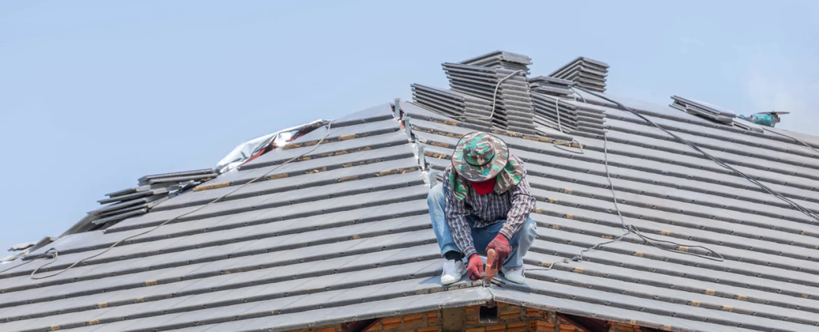 Construction worker installing gray roof tiles on a brick house under a clear blue sky.