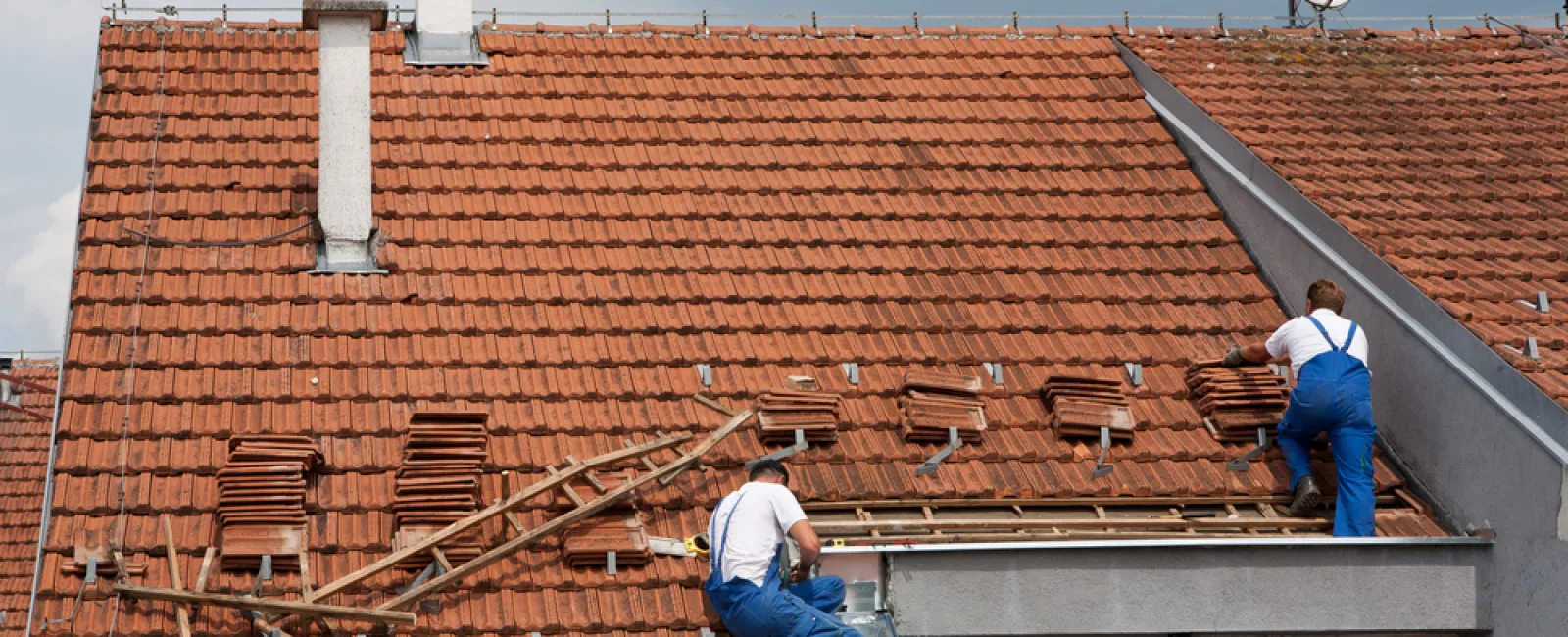 Two workers in blue overalls repairing a red tiled roof on a cloudy day with tools and stacked tiles