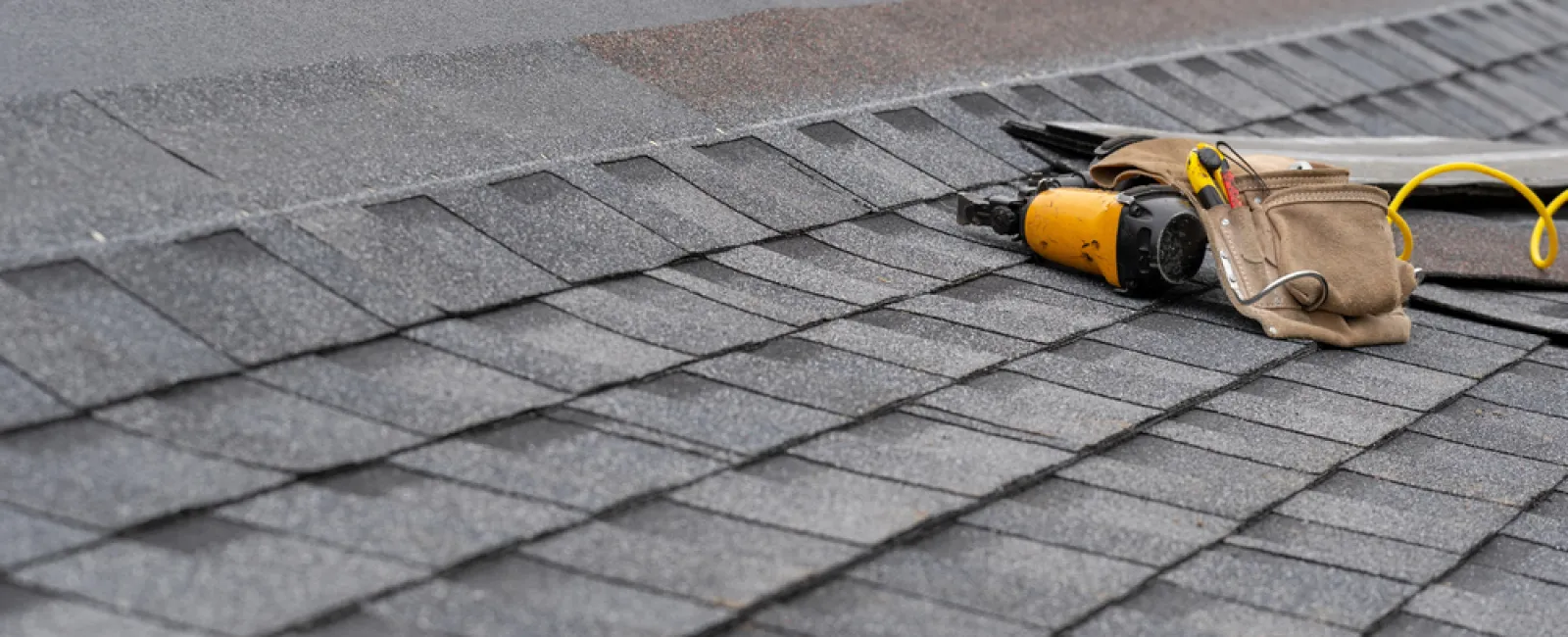 Close-up of newly installed gray asphalt roof shingles with a nail gun and tool pouch on the roof.