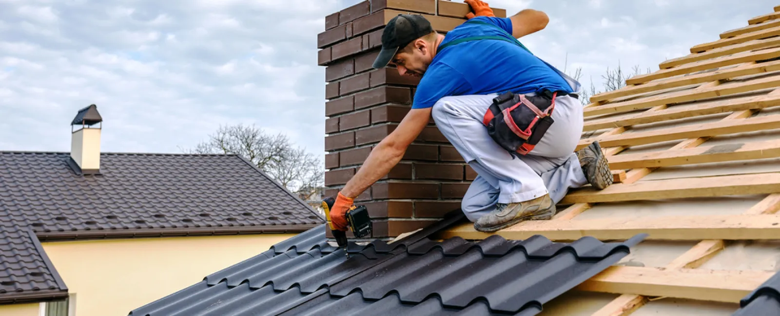 Roofer installing dark metal tiles near brick chimney on house under cloudy sky