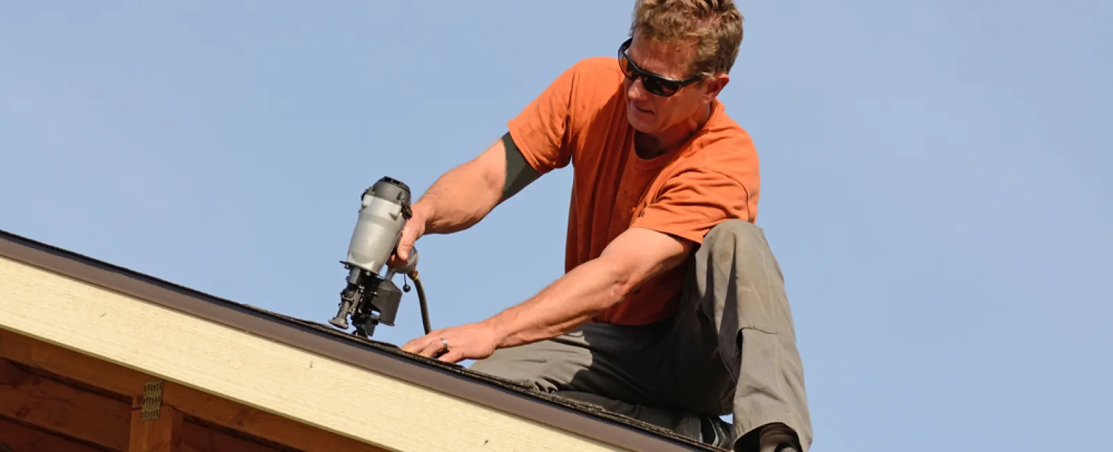 Construction worker installing roofing with nail gun on a wooden structure against a blue sky.