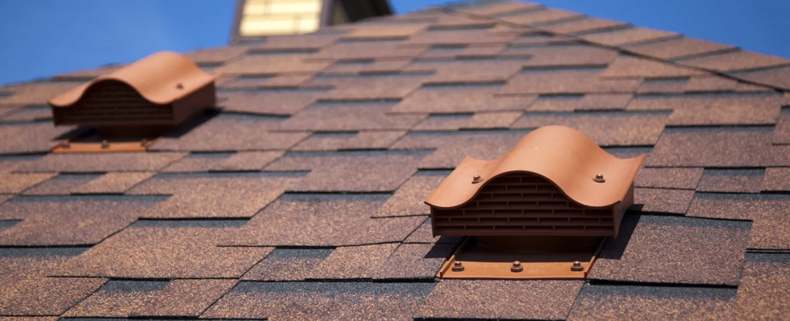 Close-up of a brown shingled roof with two terracotta-colored ventilation caps under a clear blue sky