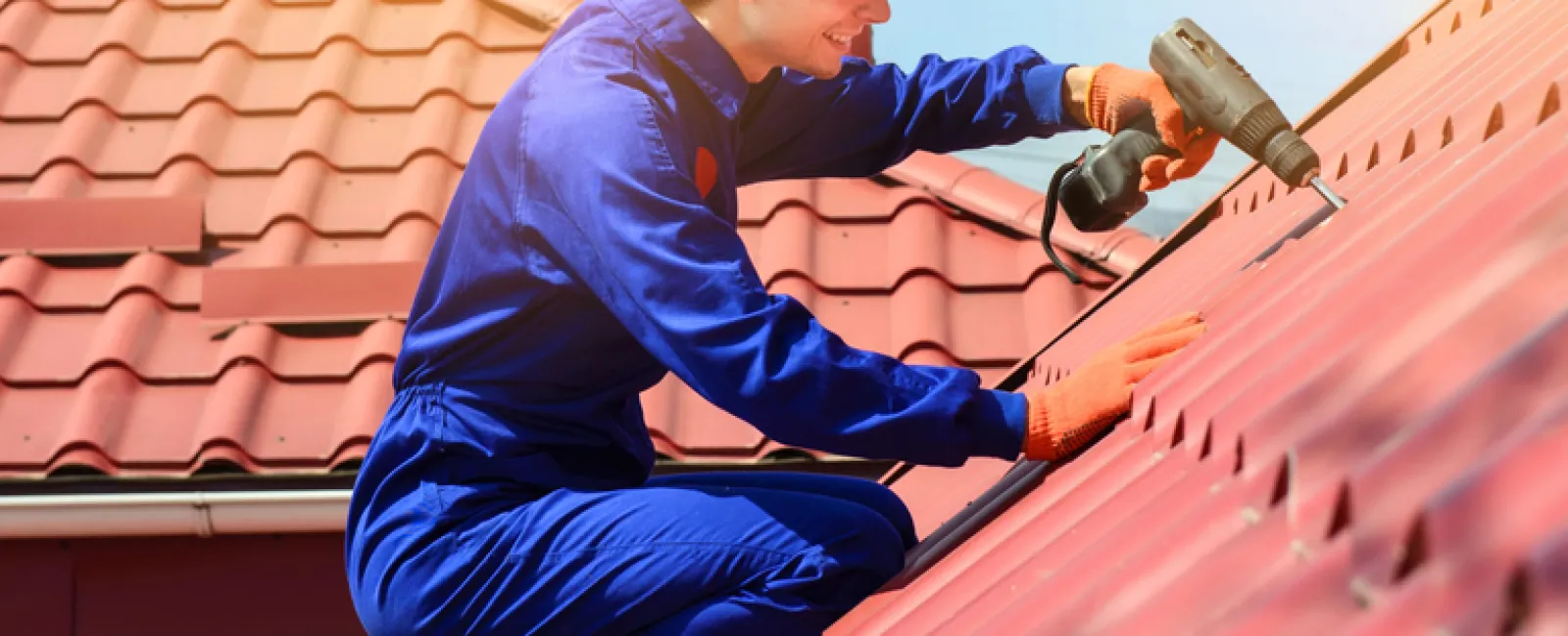 Roofing professional in blue coveralls installing red metal roof panels using a drill on a sunny day