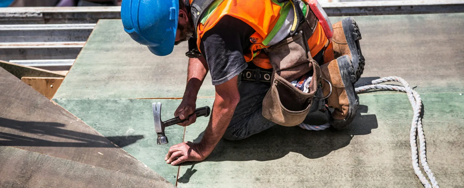 Construction worker in safety gear hammering on a roof under bright sunlight.