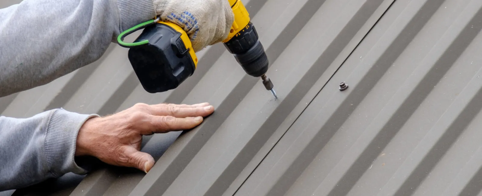 Worker installing screws on a metal roof using a yellow cordless power drill with gloved hand