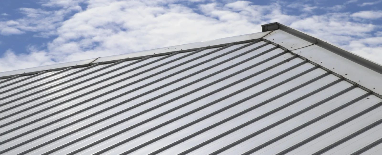 Shiny silver metal roof panels on a house under a blue sky with scattered white clouds.