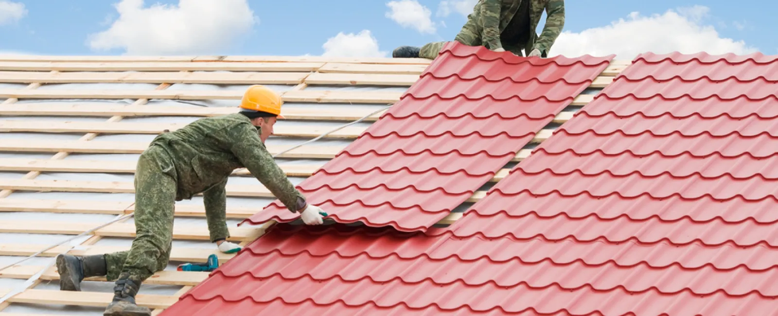 Two workers installing red metal roofing sheets on a wooden roof frame under a blue sky.