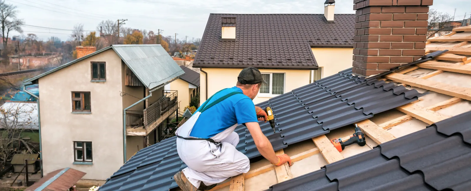 Worker installing dark gray metal roofing panels on a residential house roof on a clear day.