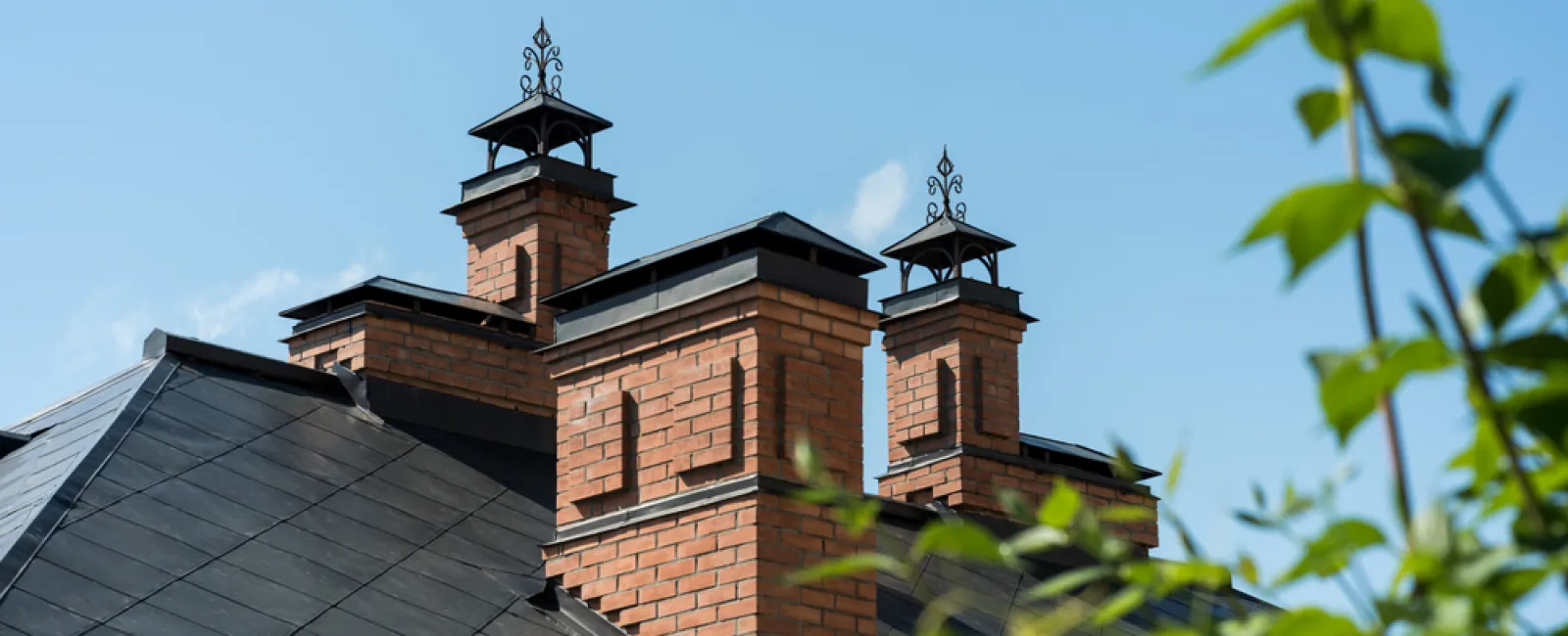 Close-up of a house roof with three brick chimneys against a clear blue sky and green foliage in foreground