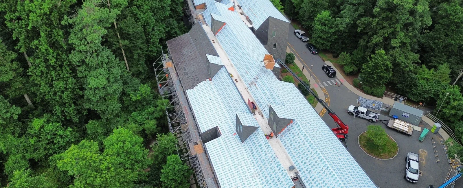 Aerial view of a large building with a newly installed white roof surrounded by dense green trees and parked vehicles.