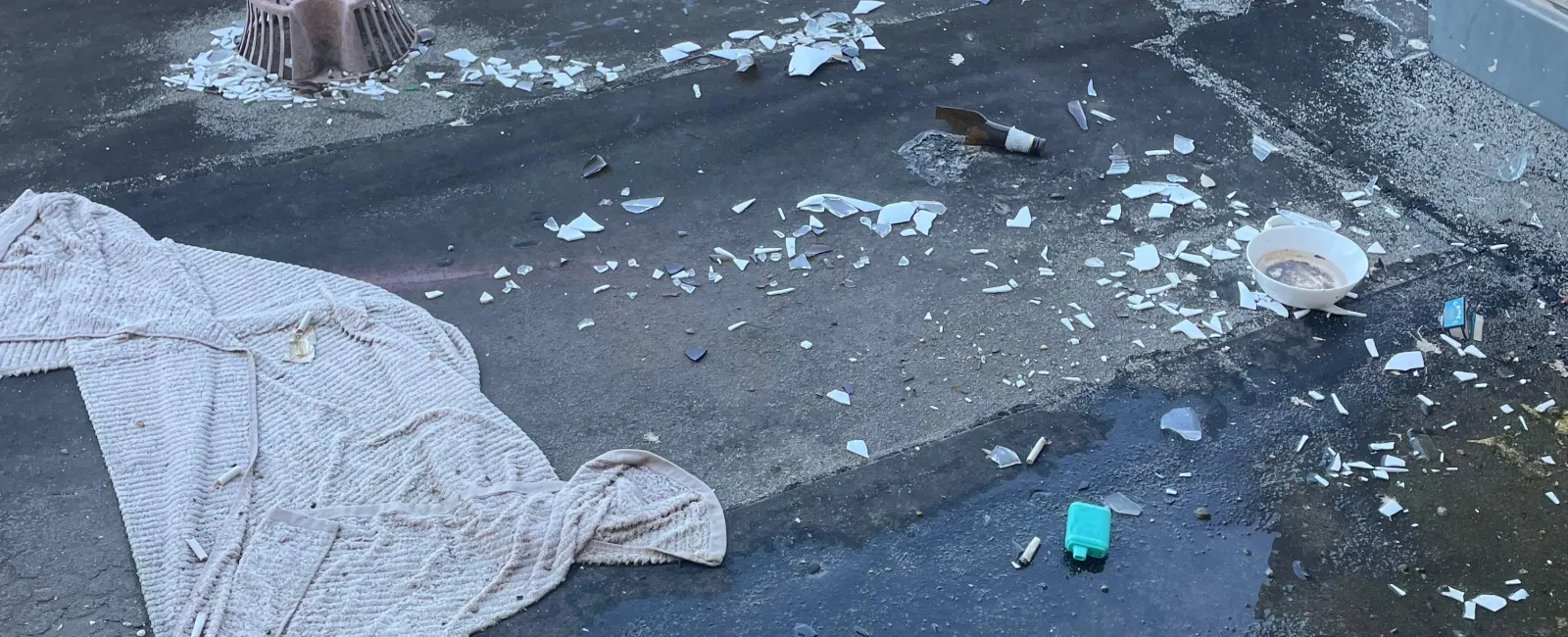 Broken glass, empty bottles, cigarette butts, and a towel scattered on a wet rooftop with greenery in background