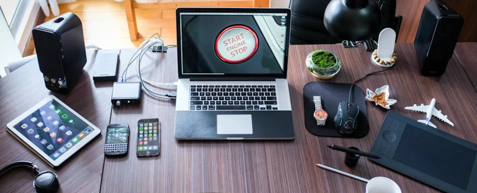 Workspace with laptop, tablets, smartphones, headphones, desk lamp, and coffee cup on wooden desk in bright room.