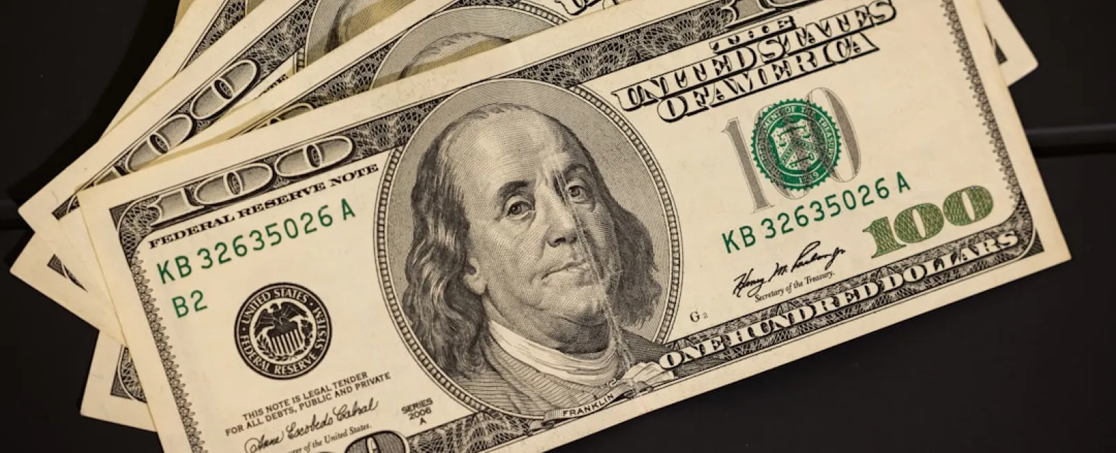 Fanned spread of US one hundred dollar bills featuring Benjamin Franklin on a dark surface.