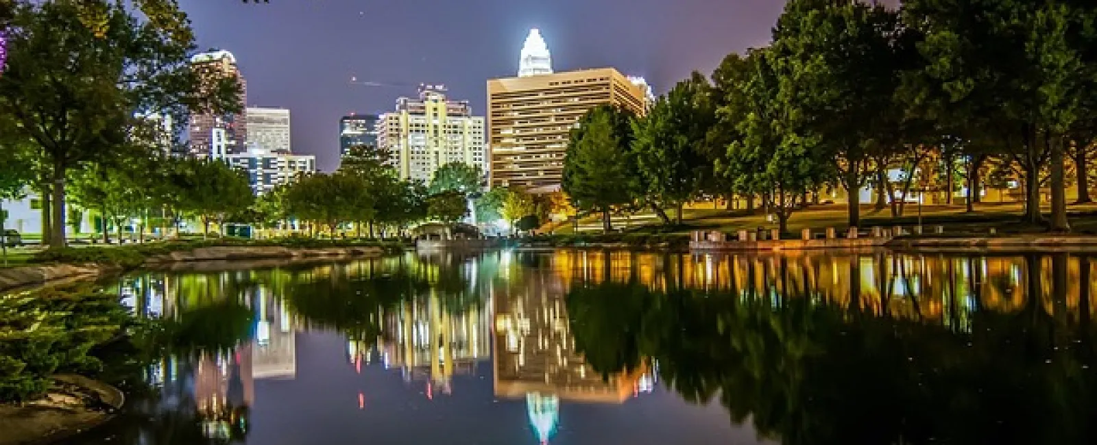 Nighttime city skyline with illuminated buildings reflected in calm river surrounded by trees and park.
