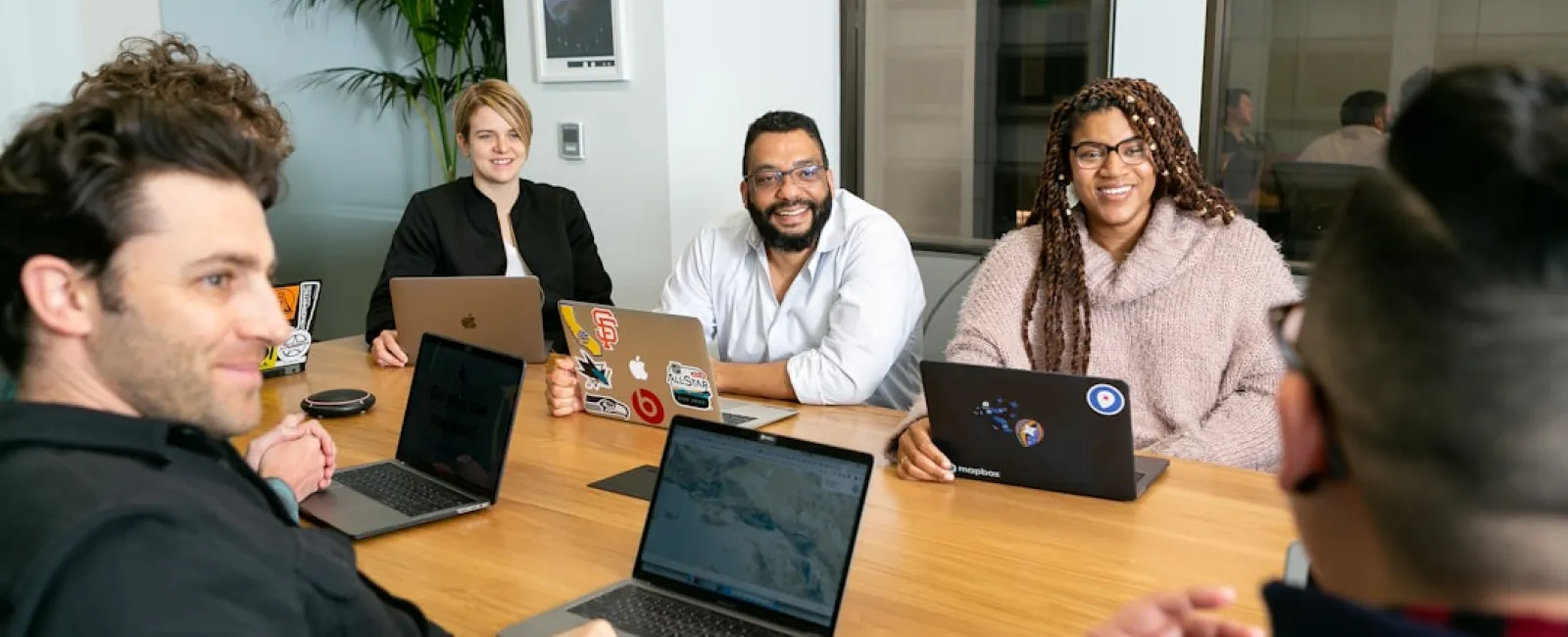Diverse group of five people in a modern office meeting around a wooden table with laptops, discussing work.