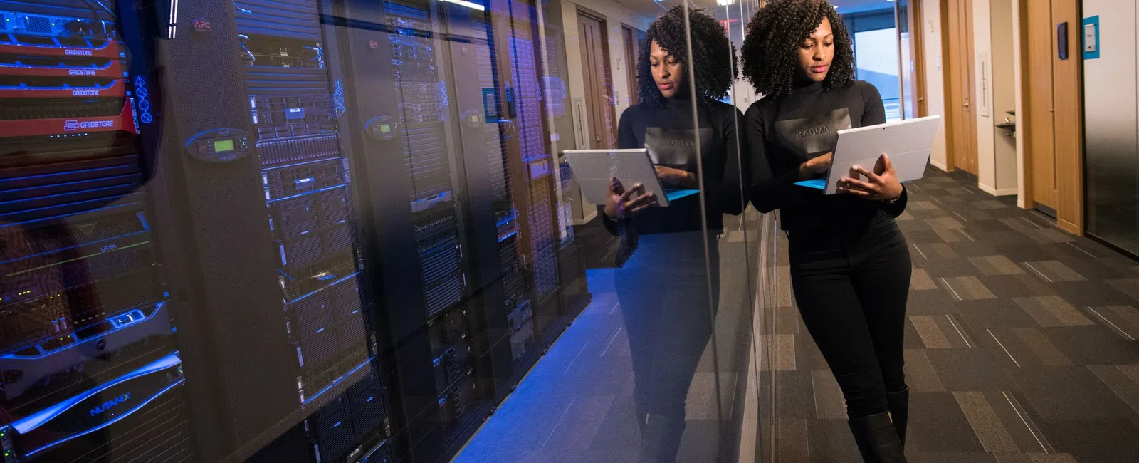 Woman in black standing in a server room hallway working on a laptop with server racks reflected on glass wall.