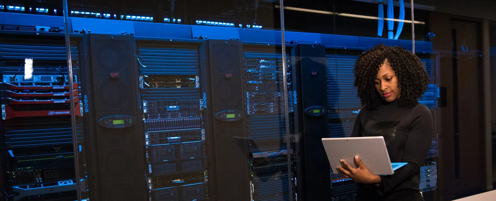 Woman standing with a laptop in a server room with multiple data racks and blue lighting.