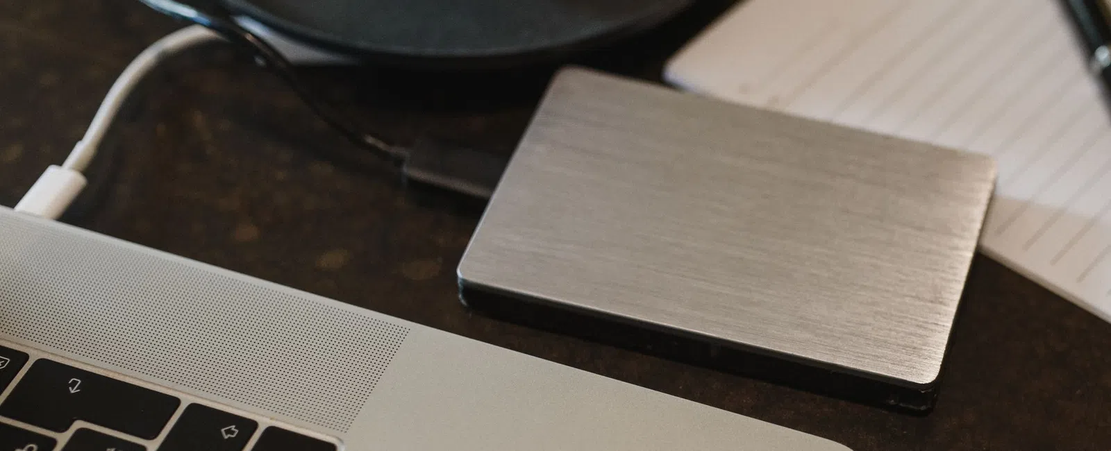 Close-up of a laptop connected to a silver external hard drive on a dark surface with a notebook and pen nearby
