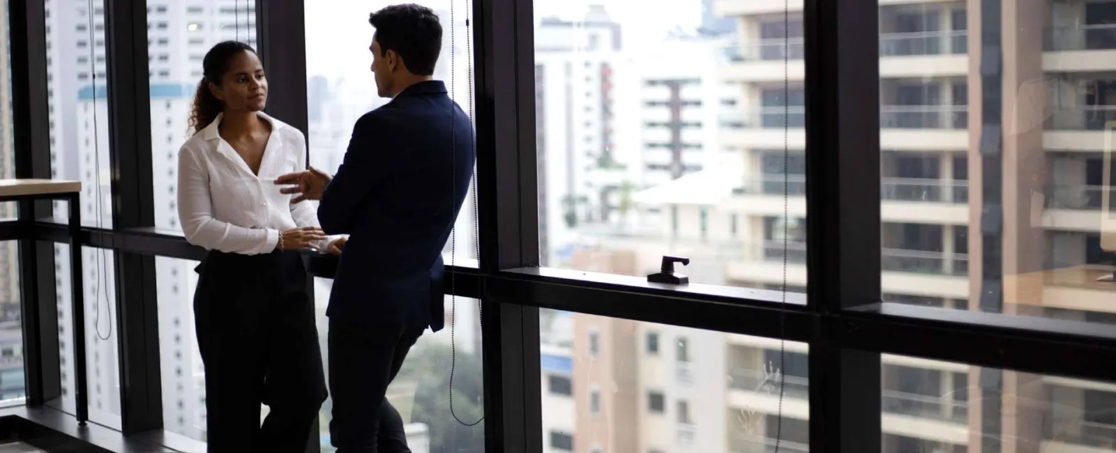 Two business professionals having a discussion near large office windows with cityscape background.