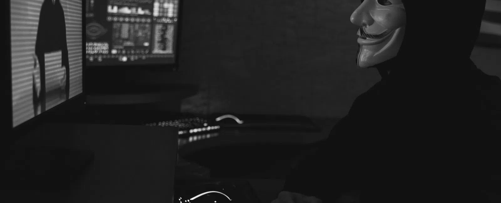 Person in Guy Fawkes mask and hoodie in dark room facing computer screen with hacker visuals.