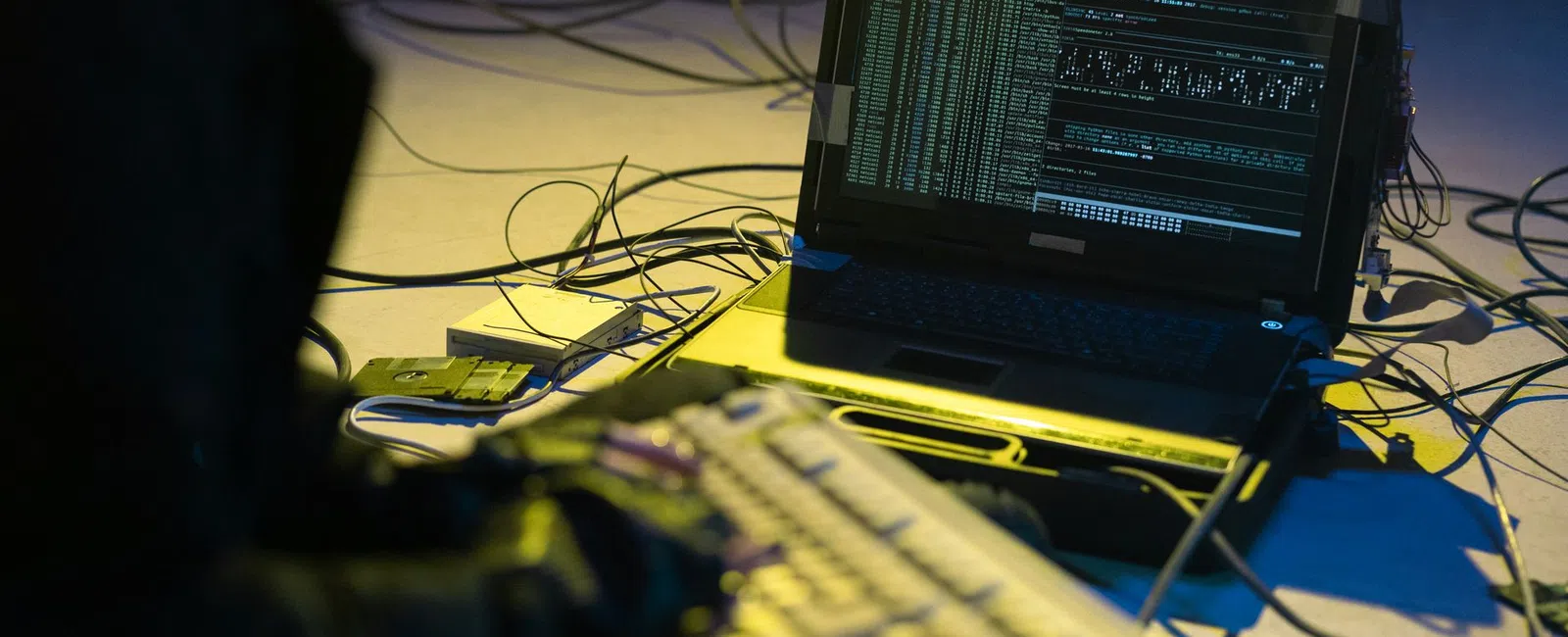 Person typing on keyboard connected to laptop displaying complex code and data with multiple cables around.