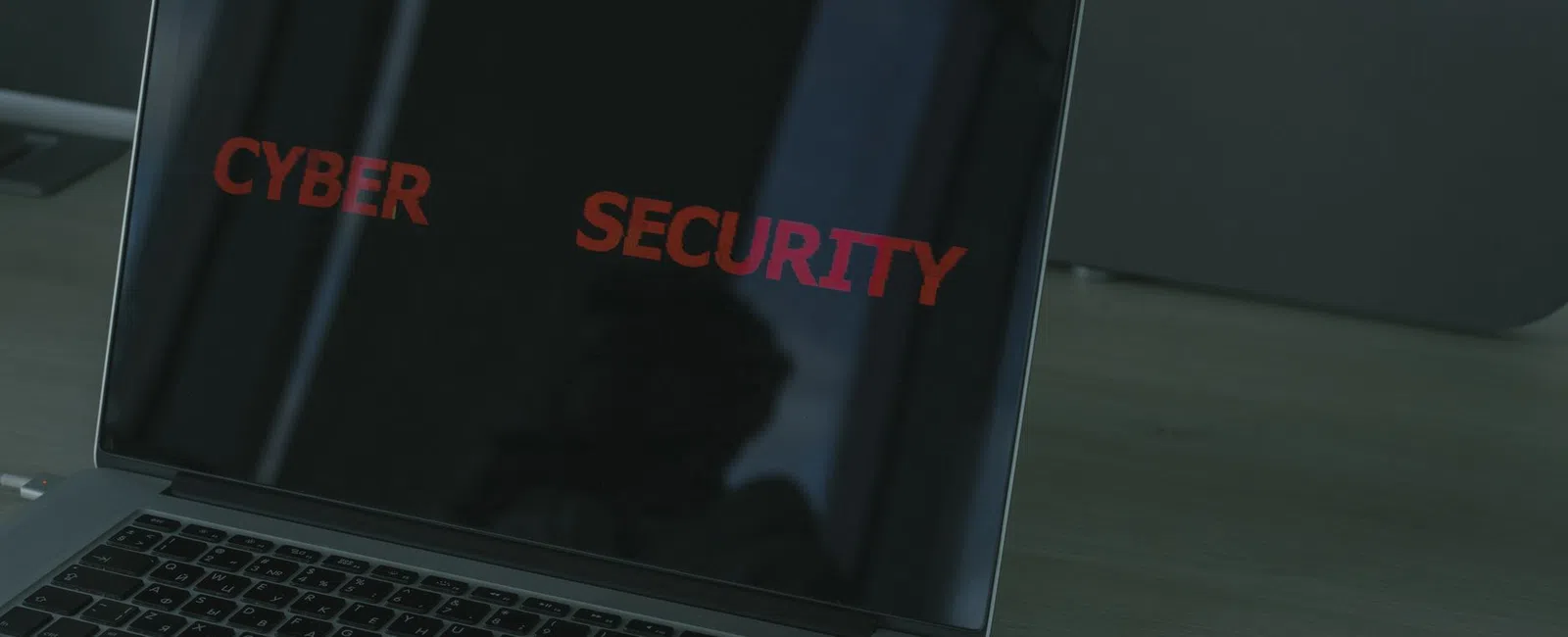 Laptop screen displaying red text 'CYBER SECURITY' on a dark background on a wooden desk.
