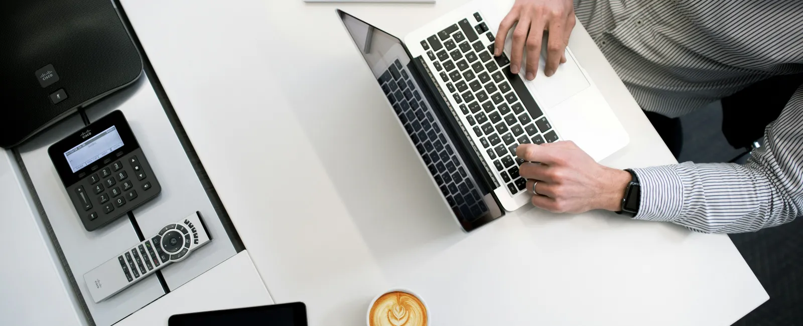 Person typing on laptop at white desk with coffee cup, tablet, phone, and notepad with pens.