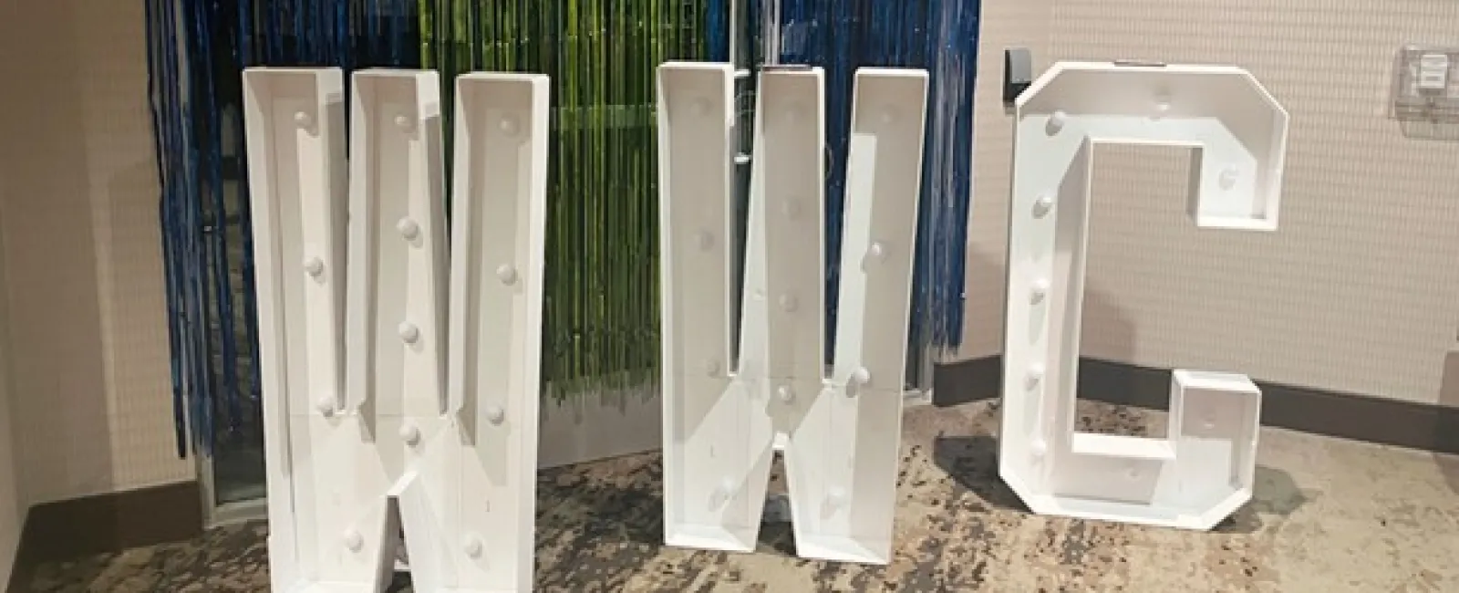 Large white marquee letters W, W, and C with light bulbs, standing on a carpeted floor against colorful decorations.
