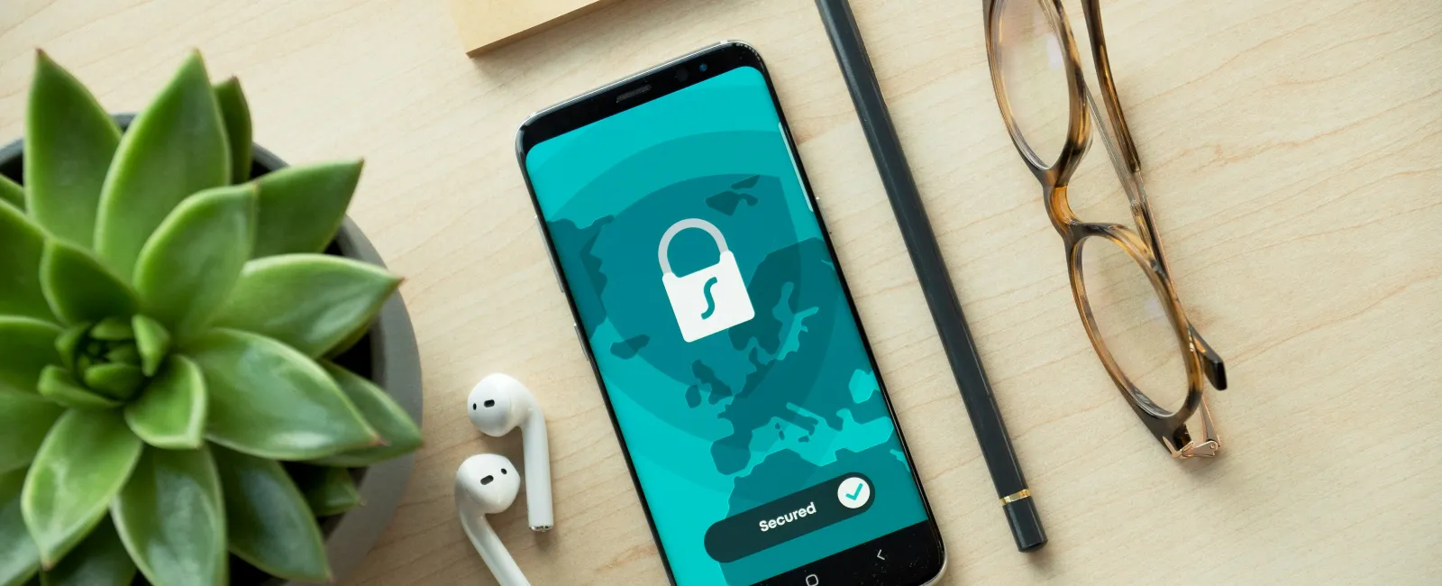 Smartphone on wooden desk showing secured lock icon, next to plant, pencil, glasses, and wireless earbuds.