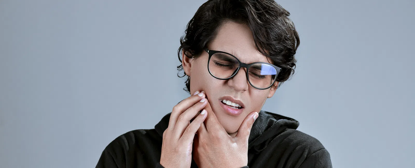 Young man with glasses in black hoodie expressing jaw pain against gray background