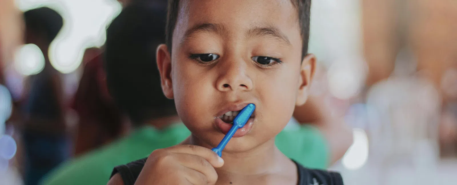 Young boy brushing his teeth indoors with a focused expression and blurred people in the background