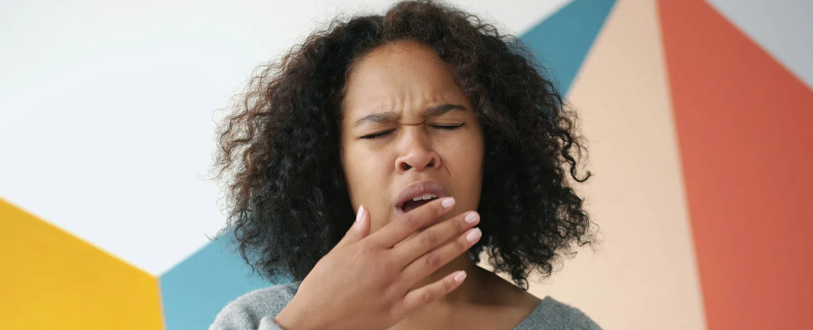 Young woman with curly hair yawning and covering her mouth wearing gray sweater against colorful wall.