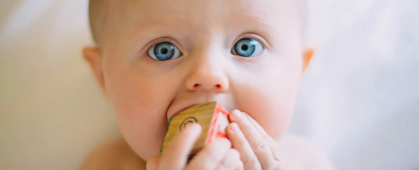 Close-up of a baby with blue eyes chewing on a colorful wooden toy block indoors