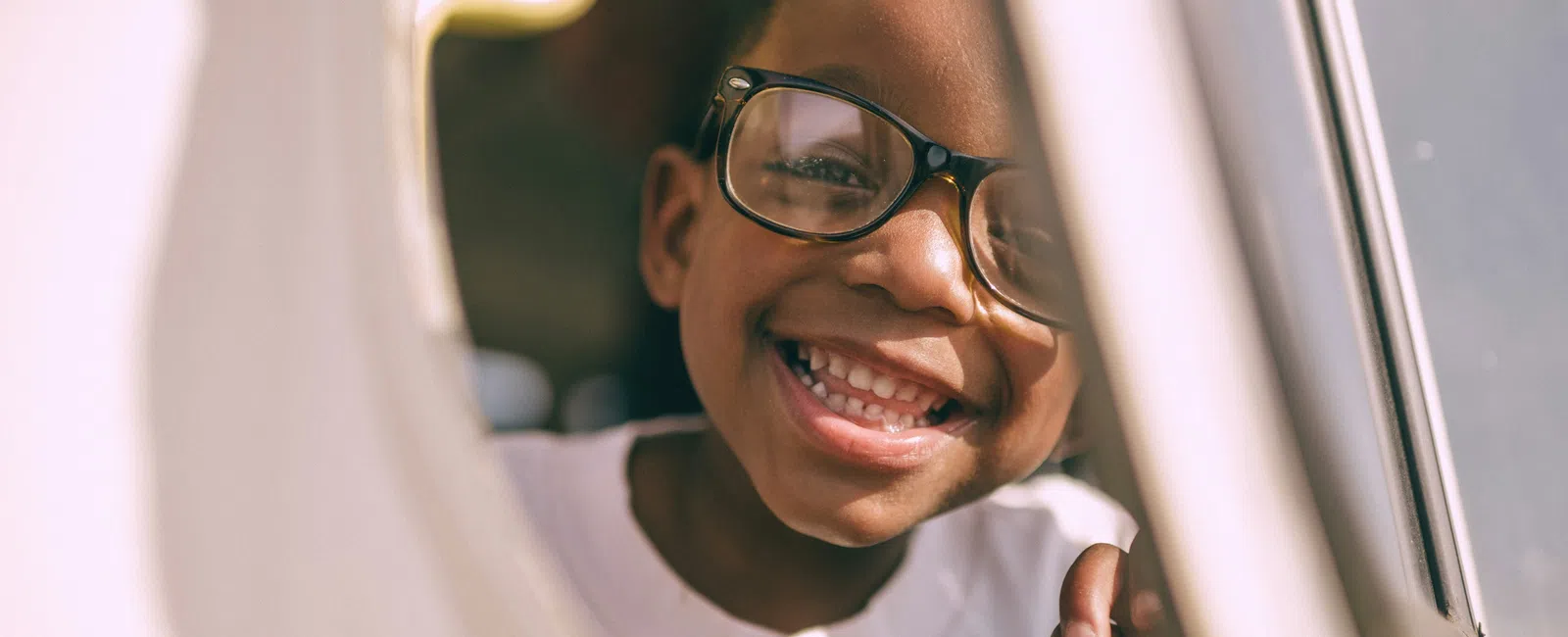 Smiling child wearing glasses looking through a round window with bright, warm lighting outdoors.