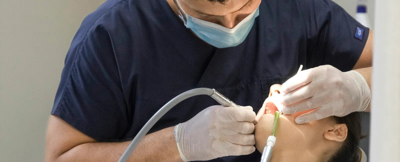 Dentist wearing mask and gloves treating patient lying back in dental chair under white light.