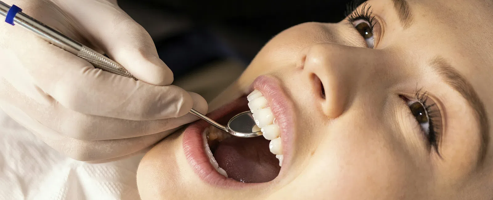 Dentist wearing gloves examines smiling woman's teeth with dental mirror during checkup appointment.