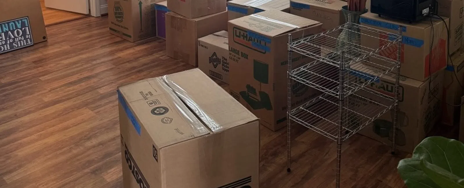 Stacked moving boxes including U-Haul, Costco, and Ace in a sunny room with wooden floor and metal rack.
