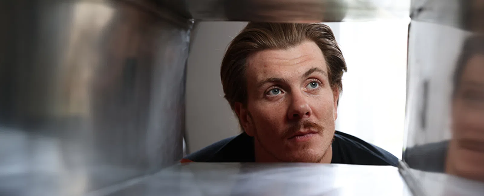Young man with mustache looking intently inside a large metallic rectangular tunnel or container from the outside.