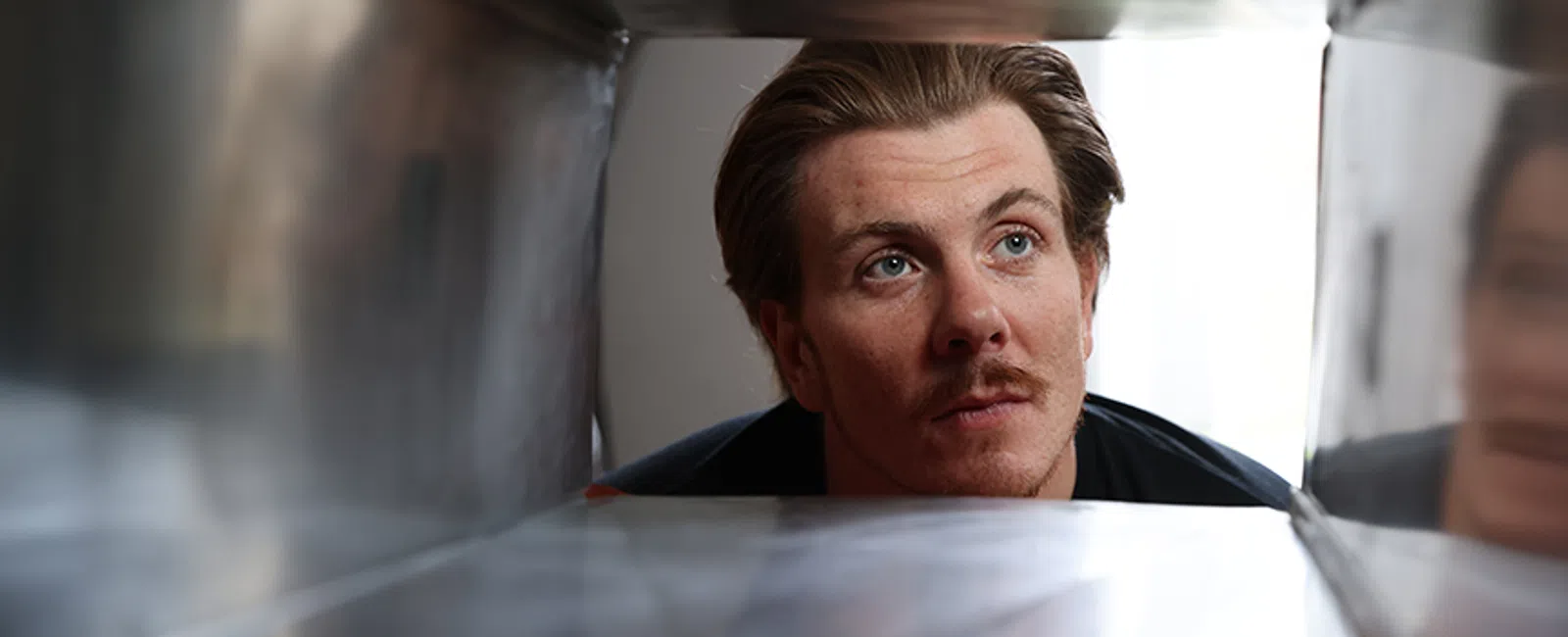 Man with mustache looking into a steel mailbox or metal container with blurred reflective interior.